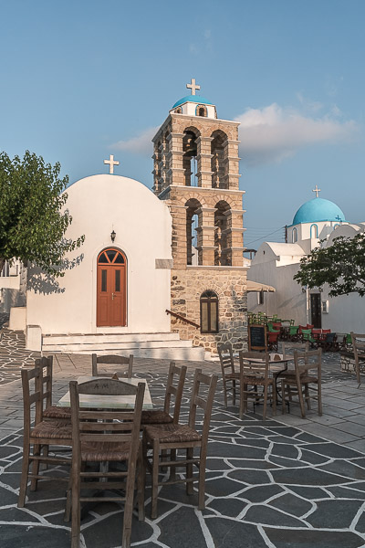 This image shows the central square of Kostos Village. There is a white church with a brown bell tower and several tables and chairs scattered around. 