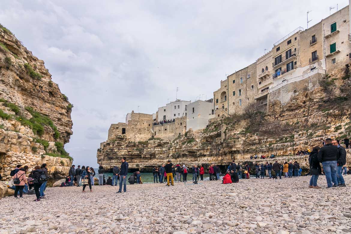 This photo was taken from Lama Monachile Beach. There are many people on the beach. Nobody's swimming because it's a cold and cloudy spring day. The Old Town looms above the beach as it sits atop a dramatic limestone cliff.
