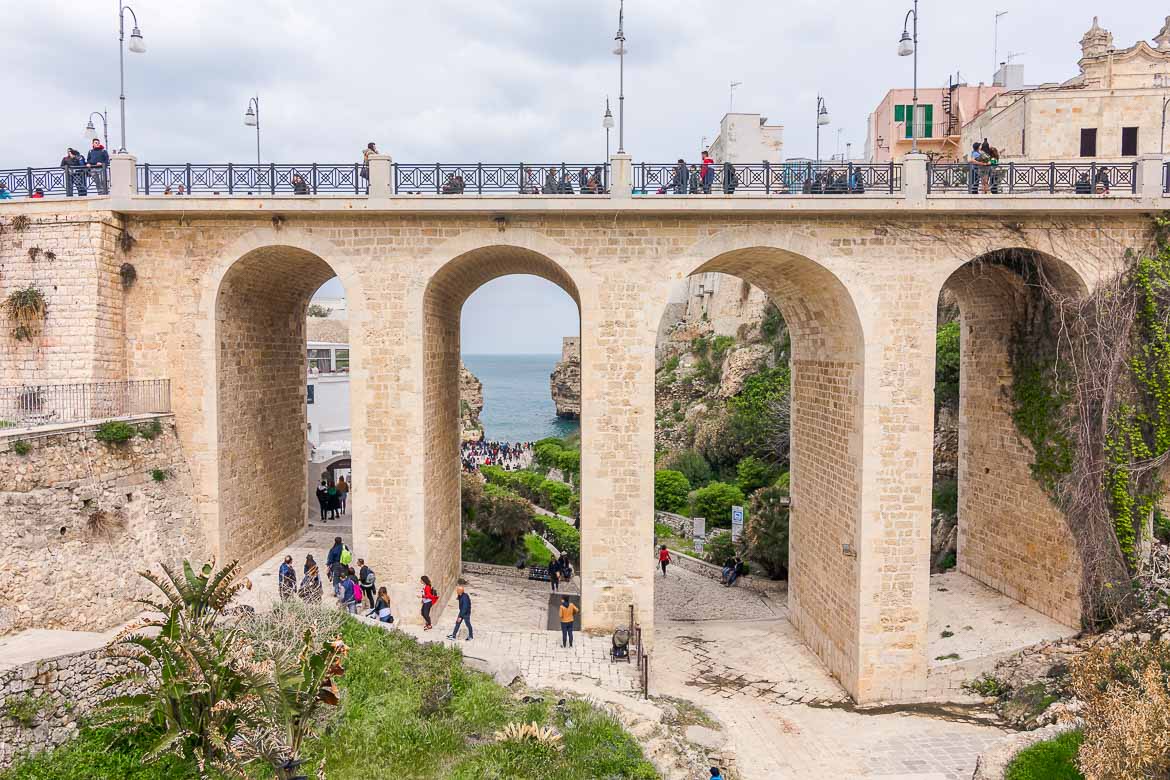 This photo shows the impressive Lama Monachile Bridge. It is a bridge with tall arches. There are people walking along the bridge. You have to walk below the bridge to get to Lama Monachile Beach. The latter is visible in the background.