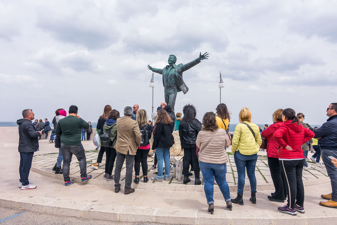 This image shows the statue of Domenico Modugno. The statue is surrounded by many people who stand to admire it.