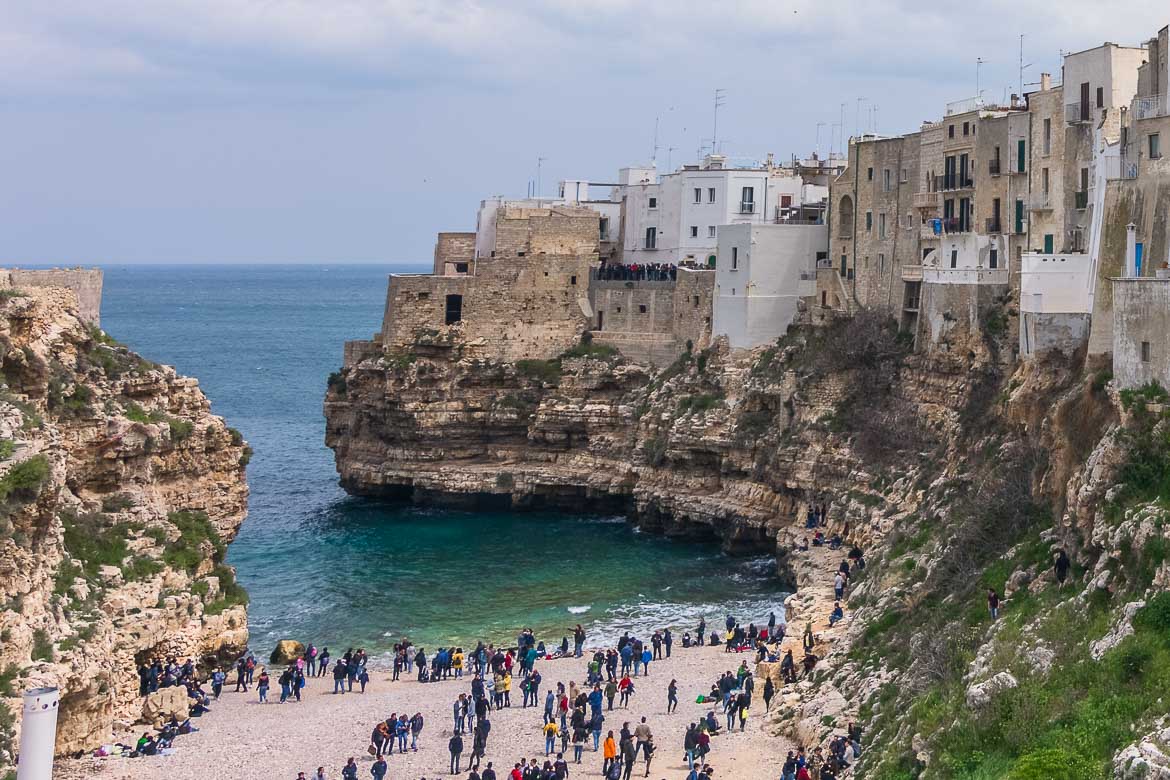 This is a panoramic view of Polignano a Mare. There are many people on the beach. The historic centre looms above in a gorgeous way.