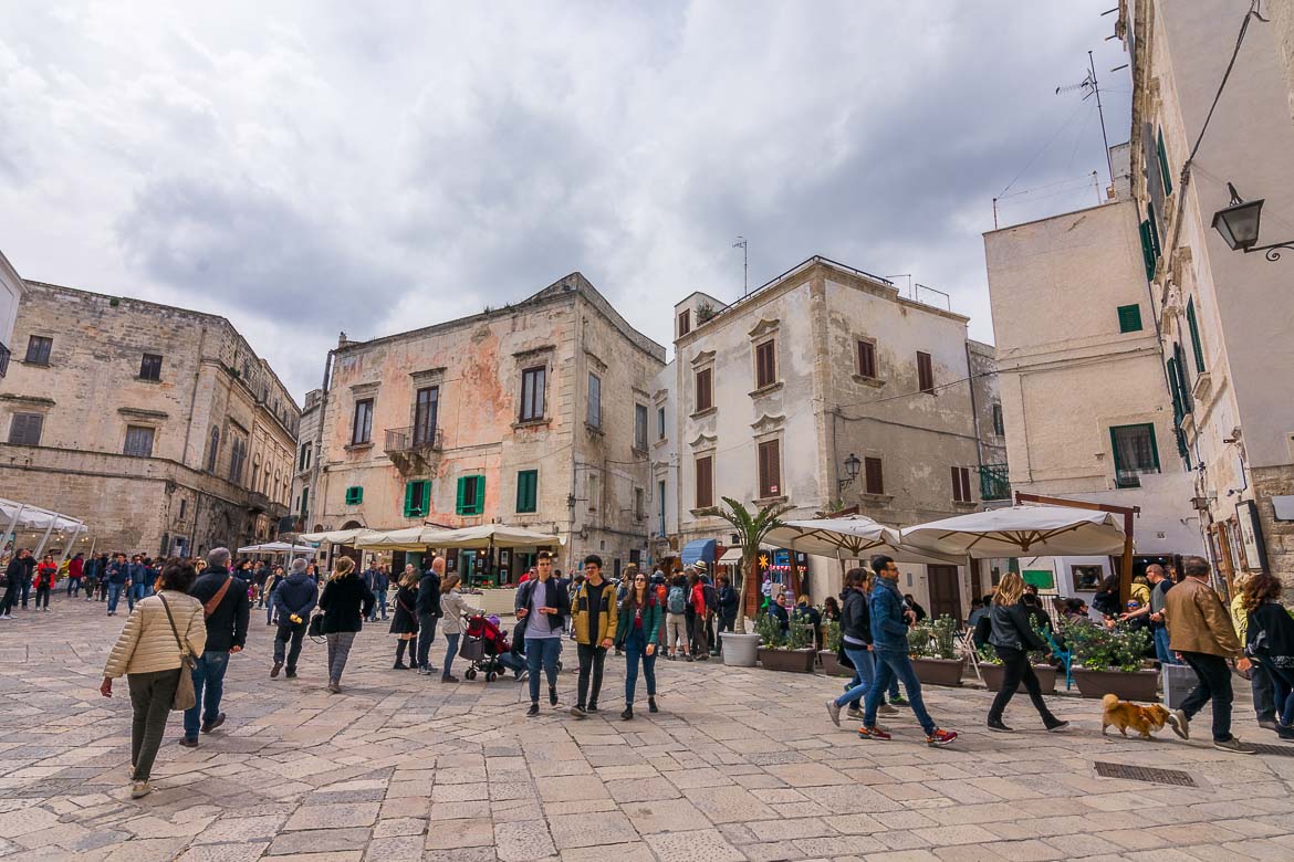 This photo shows what to do in Polignano a Mare on a busy day. People watch at Piazza Vittorio Emanuele II! The square is lined with gorgeous old buildings. There are many people walking around.