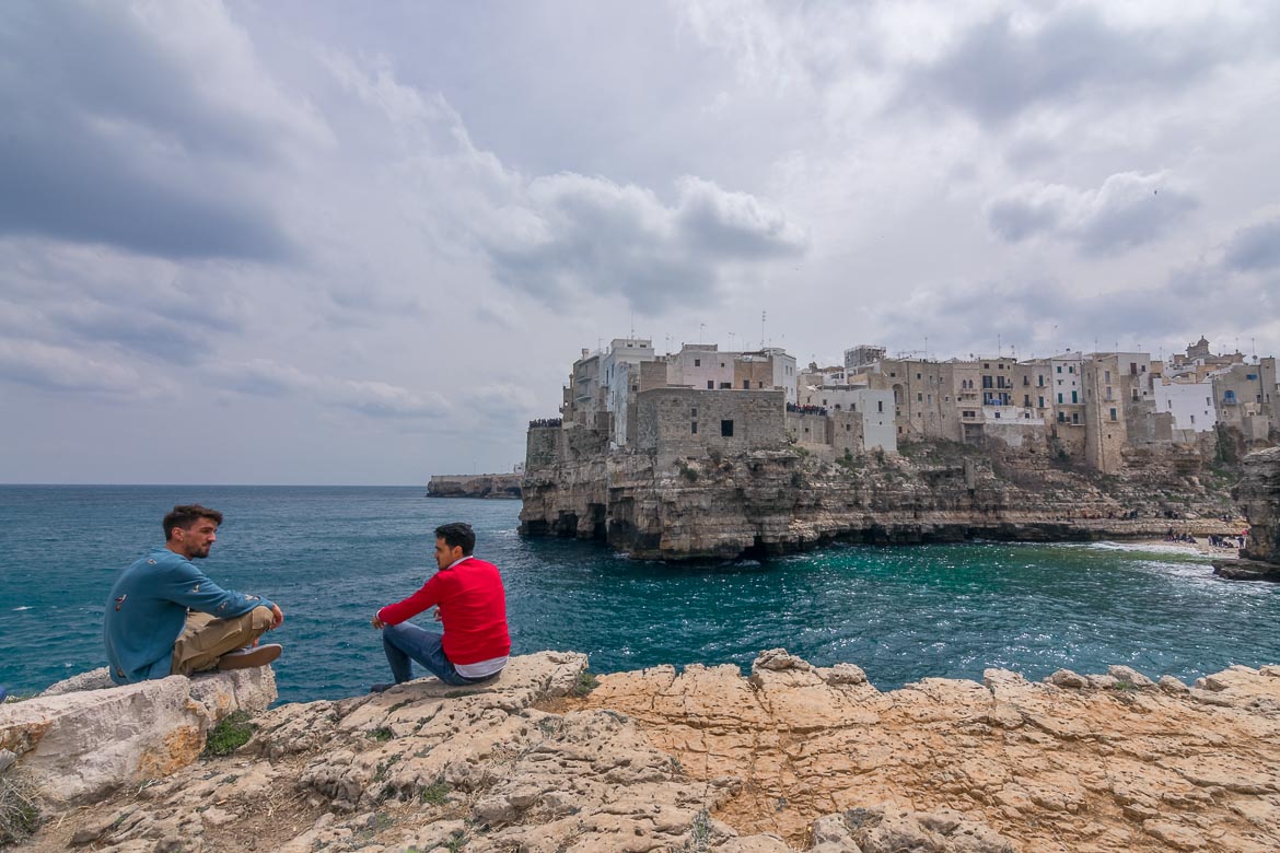 This photo shows two men sitting on the edge of Pietra Piatta, the flat rock. In the background, the gorgeous Old Town of Polignano a Mare stands atop a limestone cliff.