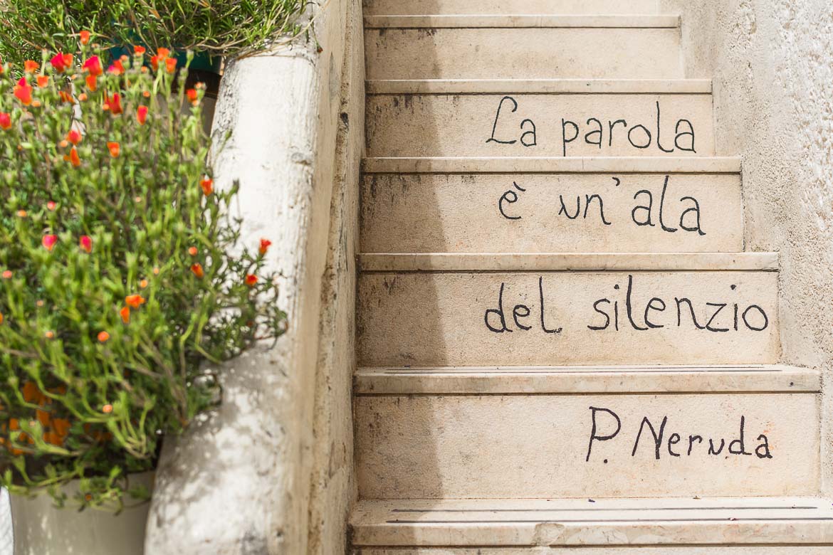 This is a photo of Poetry Steps Polignano a Mare. There's a verse from P. Neruda's Sonnet 44 written on the steps: la parola è un'ala del silenzio. This translates to: the word is a wing of silence.