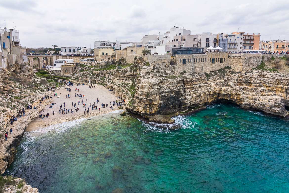 This is a panoramic view of Polignano a Mare. It was shot from Terrazza Santo Stefano, the most popular viewpoint in the town. We can see the beach with the dramatic cliffs above it. It's a cloudy spring day.