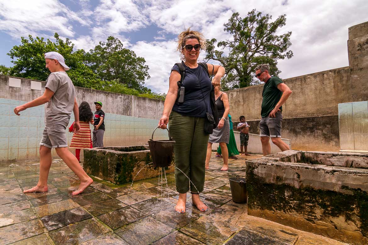 This photo shows Maria holding a bucket. The latter is dripping water from a couple of holes. Maria is smiling and happy to be playing with the water. 