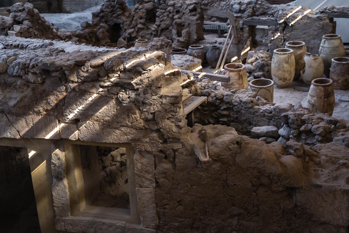 This image shows the interior of the archaeological site of Akrotiri. We can see the ruins of an ancient warehouse and several vases that have remained intact. The sunlight coming through the roof creates beautiful light effects.