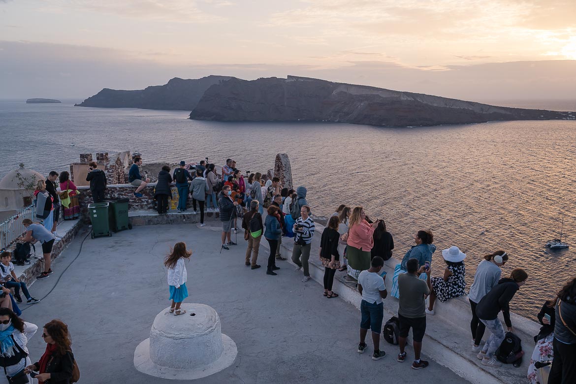 This image shows several people gathered to watch the sunset at the castle in Oia. Most - if not all - of them look at the sunset via their smartphone cameras. In the centre of the photo, there's a girl playing and dancing, probably the only person in this shot that lives the moment rather than record it.