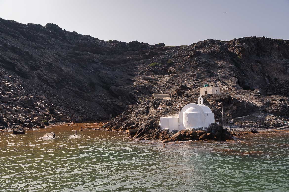 This image shows the whitewashed chapel of Agios Nikolaos, which is built on a pile of rocks in the sea. The water is green and it becomes brown closer to the shore. There are people swimming in the hot springs.