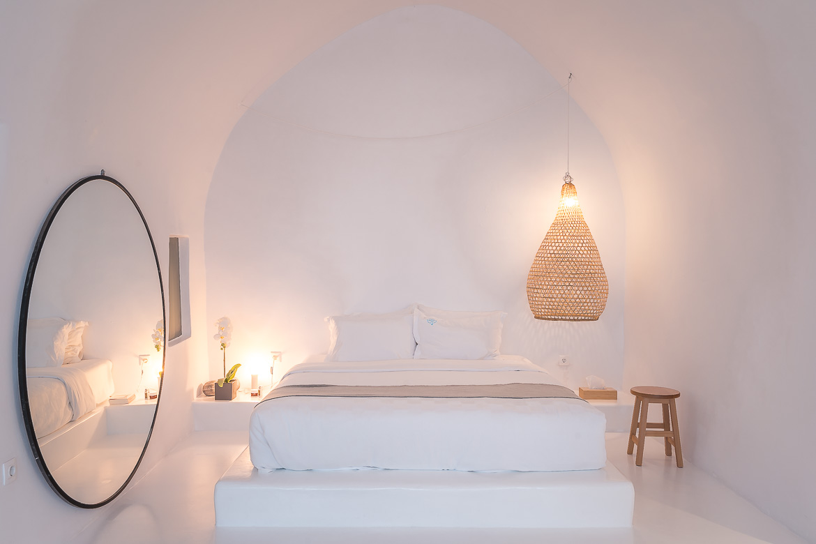 This image shows the all-white cave-like bedroom at Sophia Castle View Hotel in Oia. Apart from the bed, there's a round mirror and an impressive lamp made of straw. 