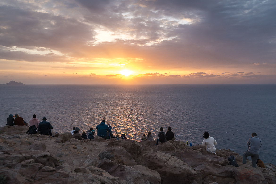 This image shows people sitting on the rocks, watching the sun dive into the sea. The photo was taken from Akrotiri Lighthouse, one of the most romantic places to visit in Santorini in 4 days.