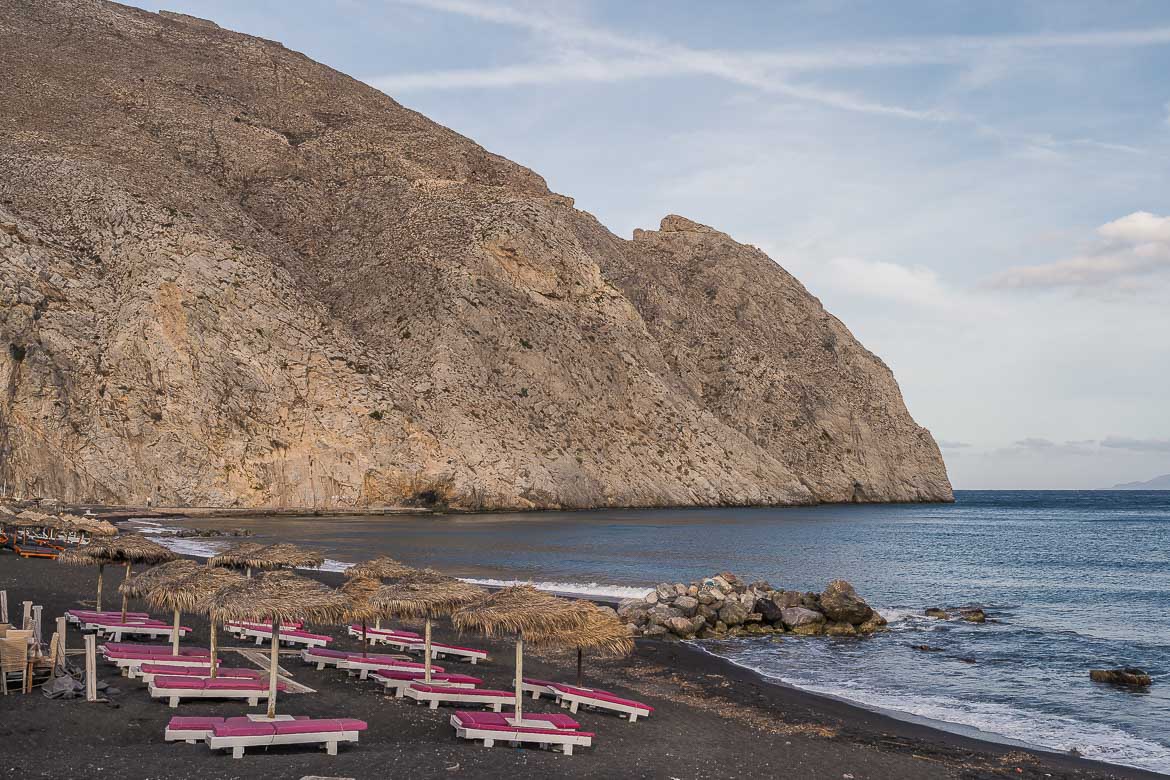 This image shows the far left end of Perissa Beach with its black sand and imposing white rock. There are sunbeds and umbrellas but no people around.