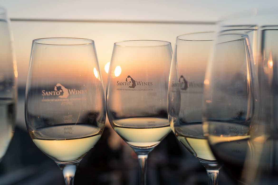 This is a close-up of several glasses of wine that bear the Santo Wines logo. Through them, we can see the sunset in the background.